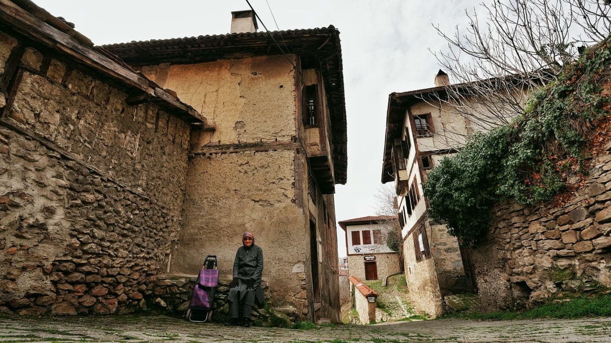 An elderly woman sits on a bench in the historic streets of Safranbolu, Türkiye, showcasing traditional architecture.