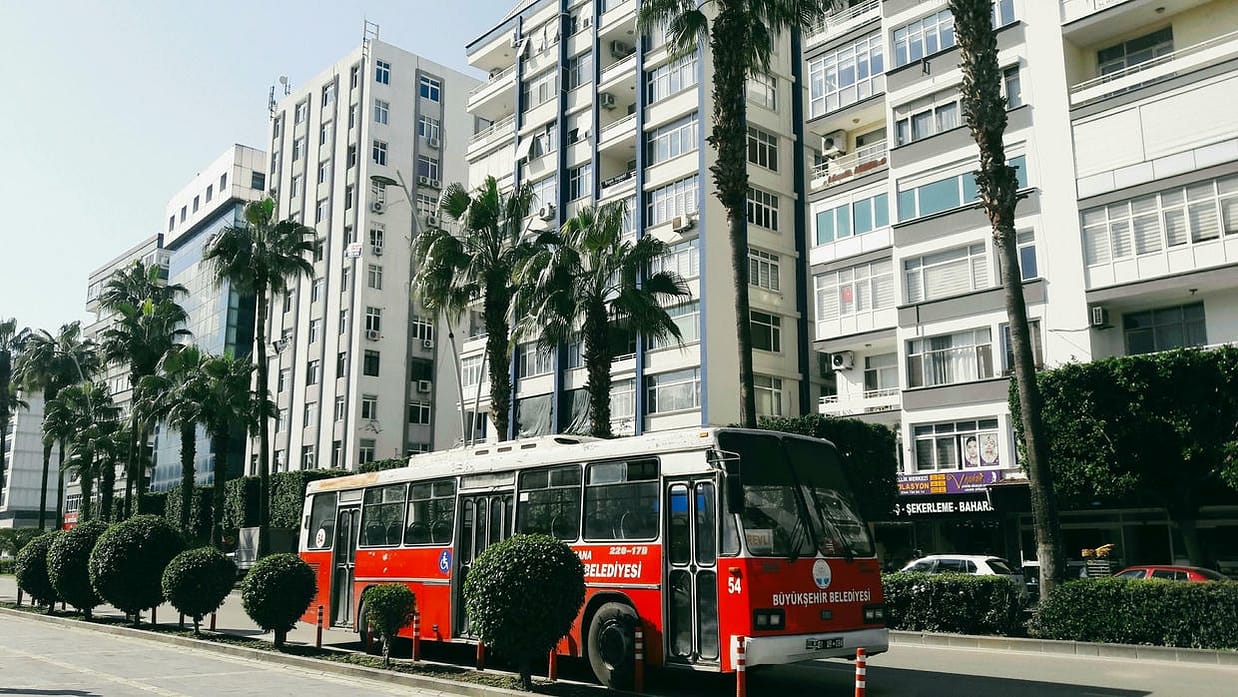 A city bus on a sunny street in Adana, Türkiye, with palm trees and urban buildings.